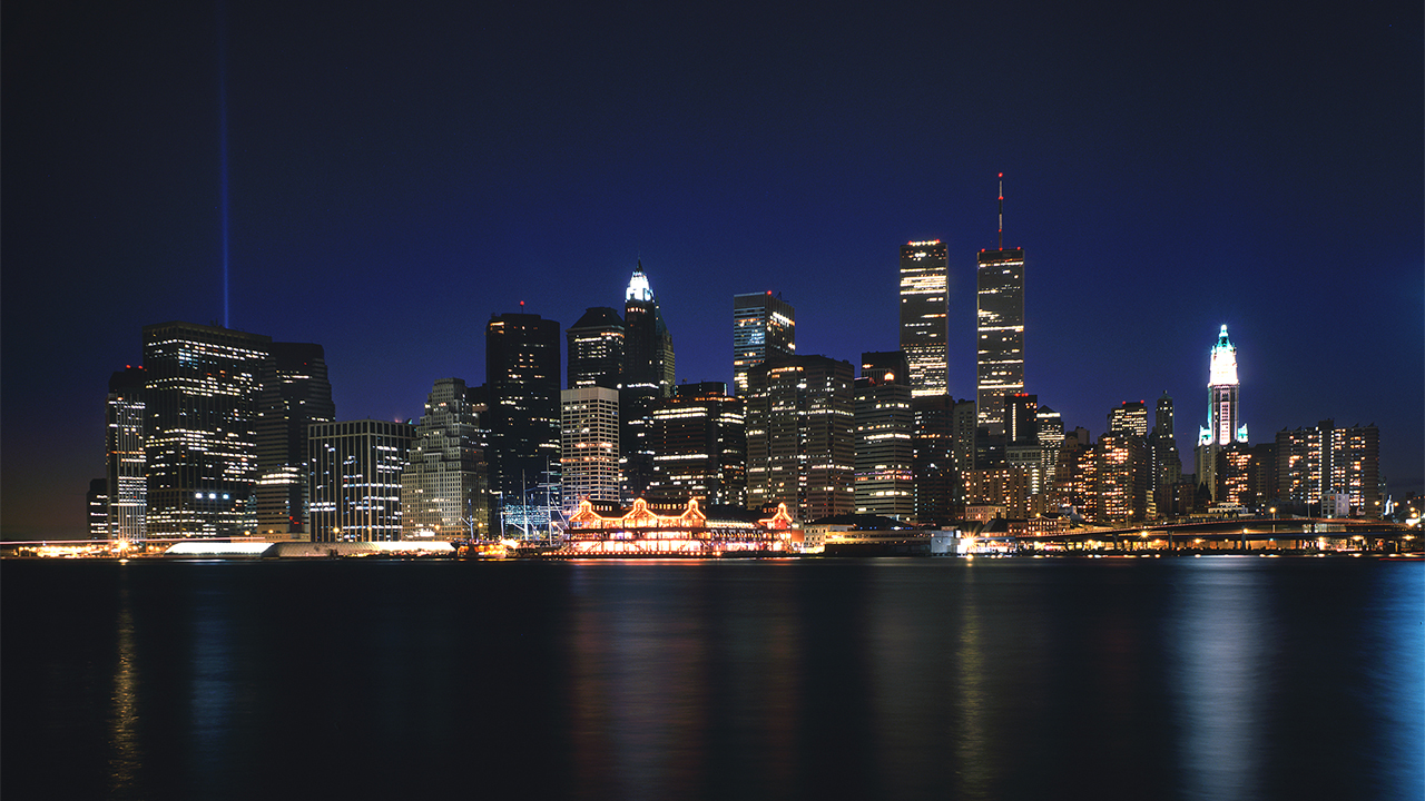 The Twin Towers stand over the rest of Lower Manhattan at night. The light of buildings reflects off the East River as the last rays of sunlight create a blue hue over the skyline.