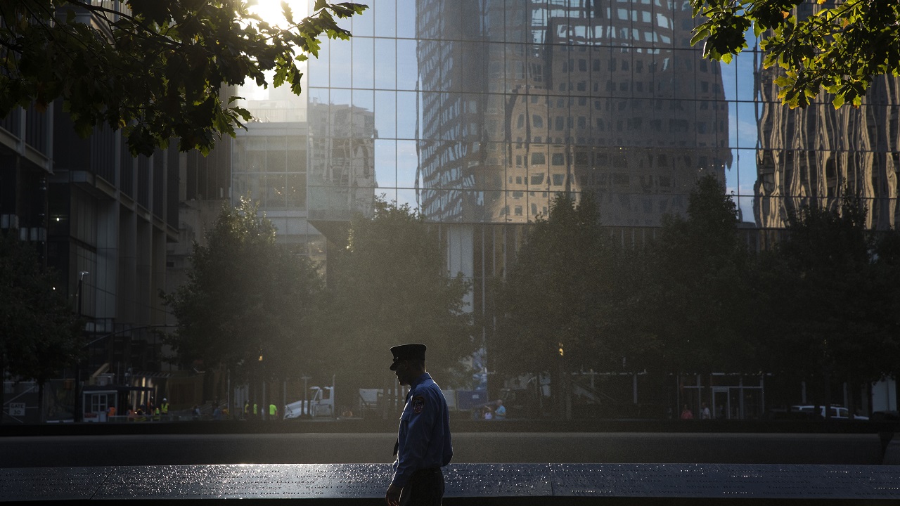 A firefighter in a formal outfit and hat looks at victims’ names on a bronze parapet. Rays of sunlight come through a gap in buildings and shine down on him and a reflecting pool. In the darkened distance are trees and building facades.