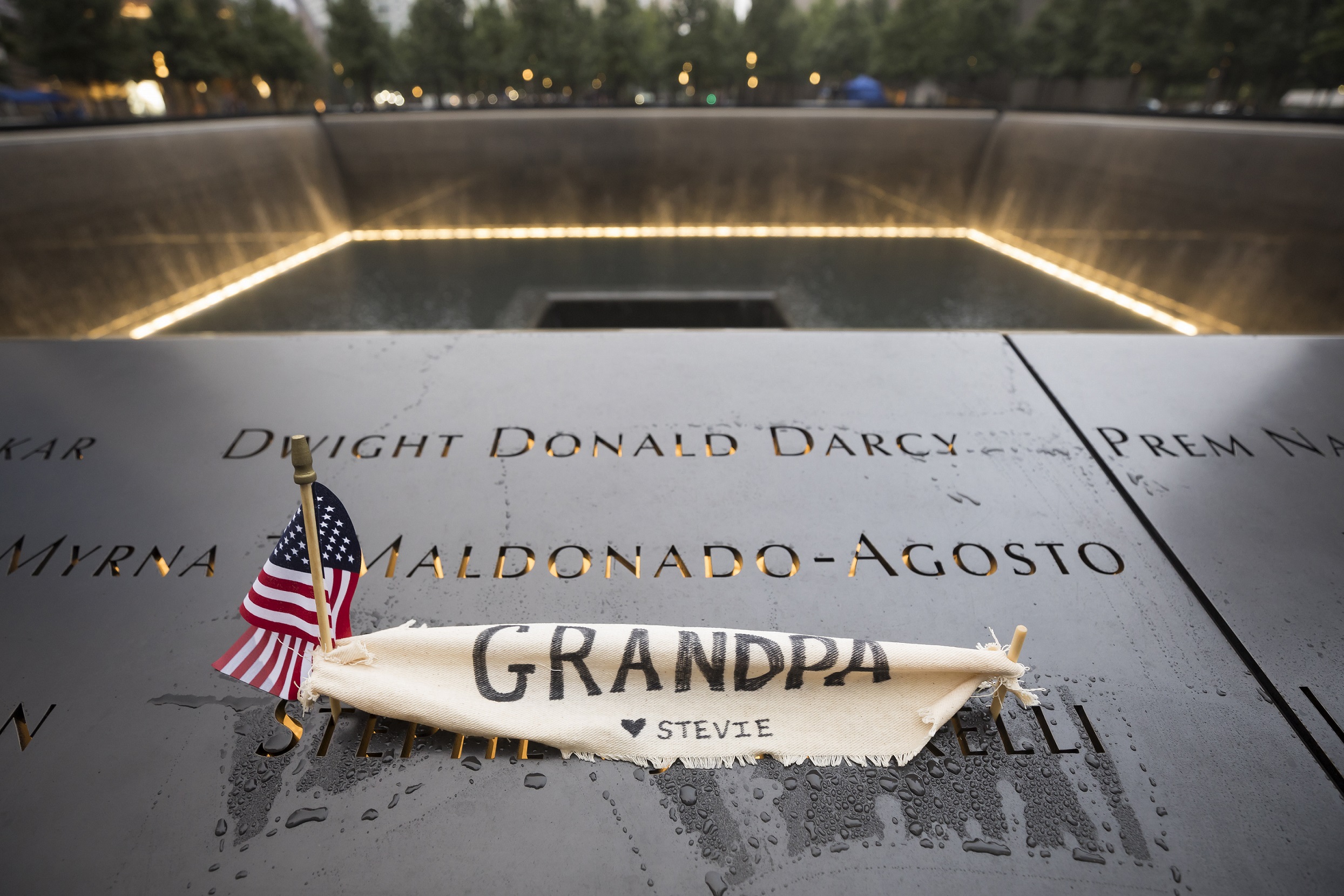 A small American flag and a hand-written note have been placed at a victim’s name on a bronze parapet at the Memorial. The note is written on a piece of cloth and reads “Grandpa.” Water cascades down a lit reflecting pool in the background.