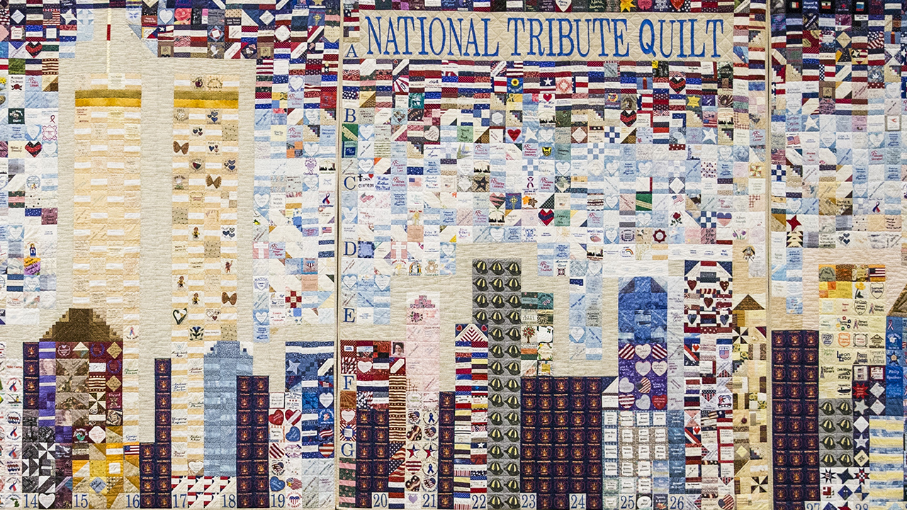 A mosaic quilt forms the Twin Towers and other buildings on the New York skyline. The quilt is made from many smaller patches that include hearts, stars, and flags from around the world. The upper right corner of the quilt reads “National Tribute Quilt.”