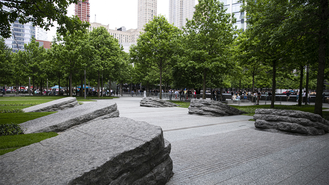 Six stone monoliths border the pathway of the 9/11 Memorial Glade. There are three monoliths on each side of the path. Trees with bright green leaves stand to the left and right of the path.