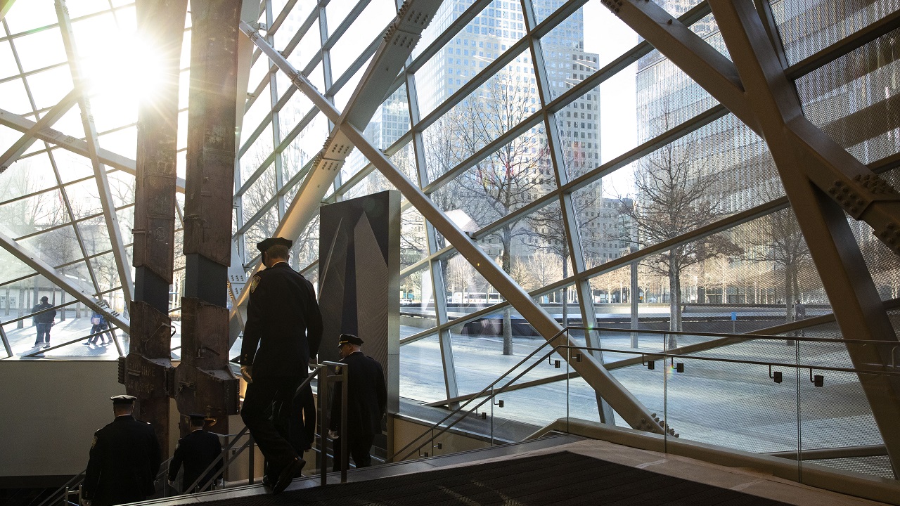 Formally dressed police officers walk down the steps to the underground 9/11 Memorial Museum as sunbeams shine through the glass of the Museum pavilion. 