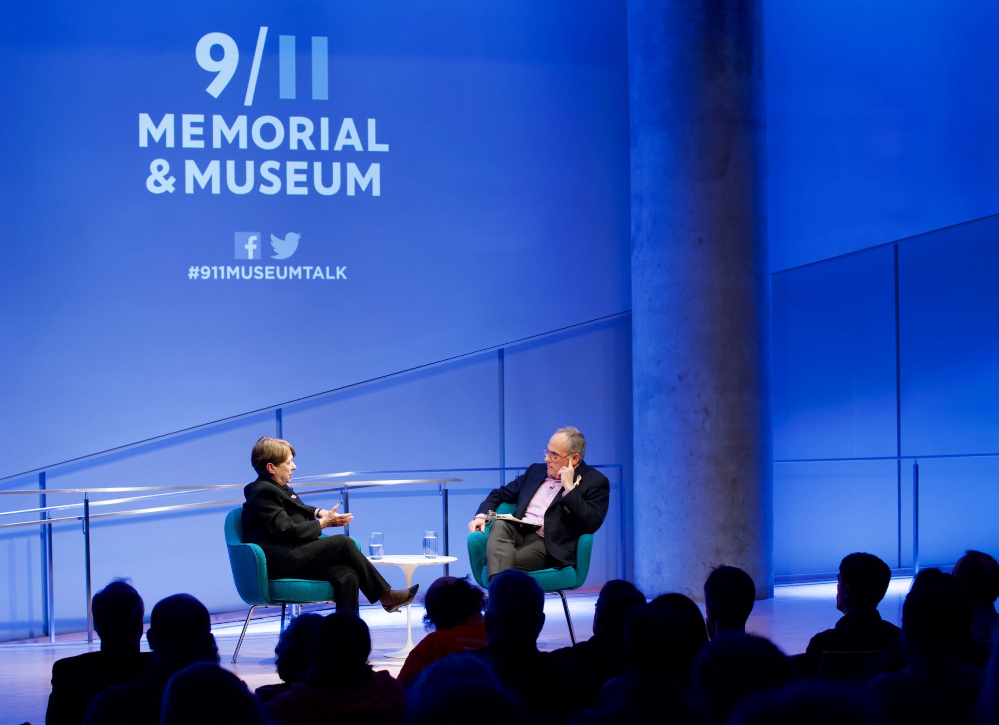This wide-angle photo shows former U.S. Attorney Mary Jo White of the Southern District of New York speaking onstage as she takes part in the public program, Prosecuting the 1993 Bombers. Clifford Chanin, the executive vice president and deputy director for museum programs, sits next to her. Members of the audience are silhouetted by the lights onstage. The logo of the 9/11 Memorial & Museum is projected on the wall above White.