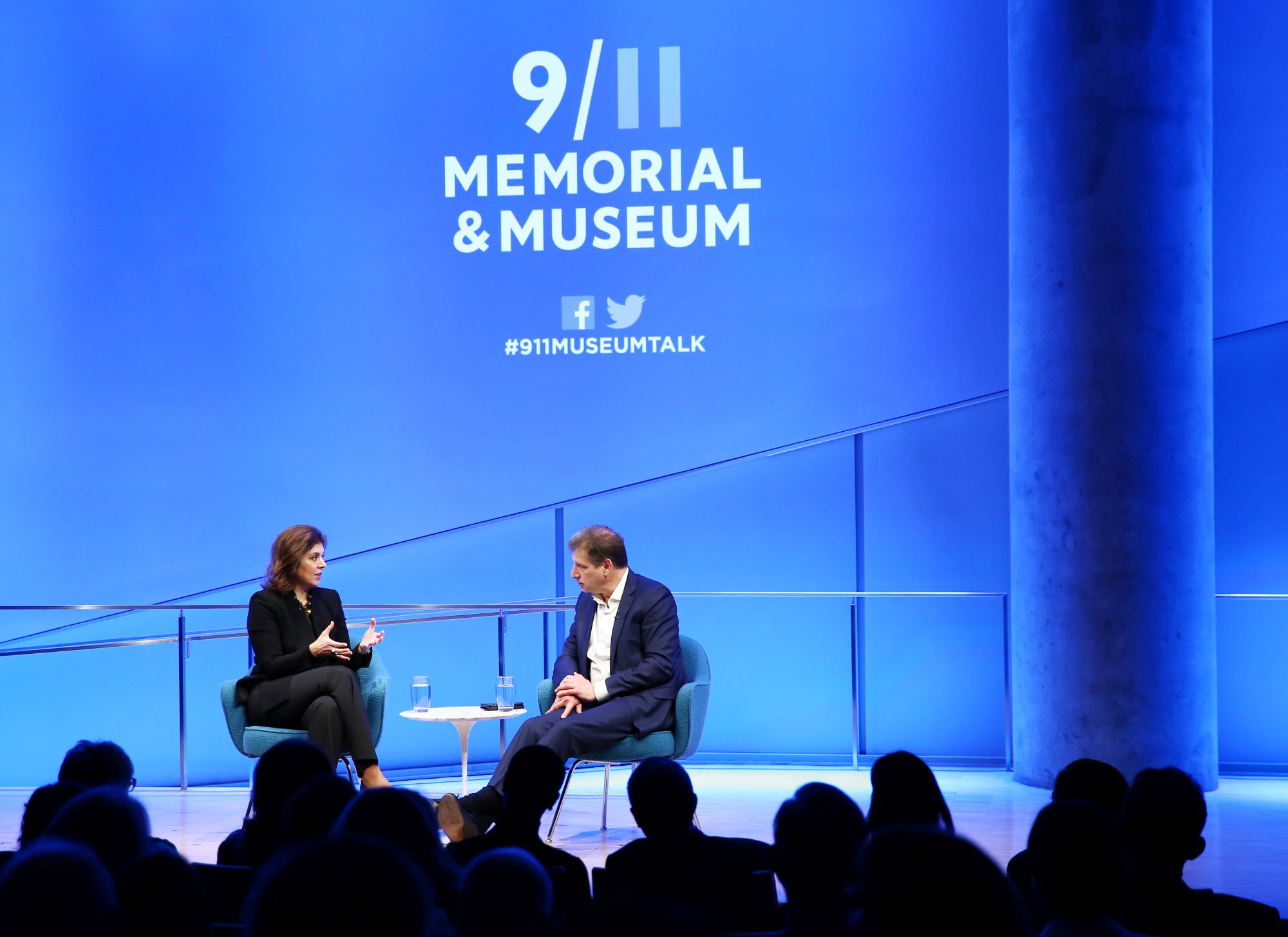 Author Farah Pandith gestures as she speaks with Foreign Affairs editor Gideon Rose onstage at the Museum auditorium. The silhouettes of audience members are visible in the foreground. The stage is lit up blue and white and the logo for the 9/11 Memorial & Museum is projected above Pandith and Rose. Audience members are silhouetted in the foreground.