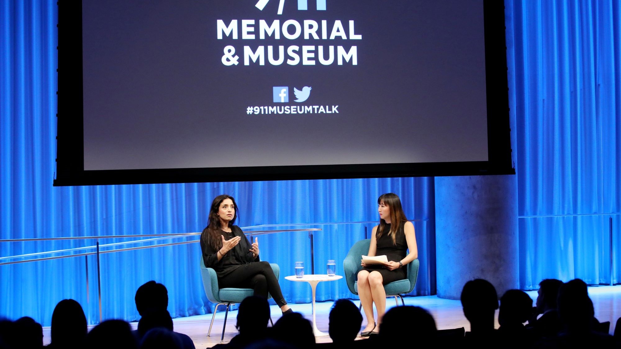 Emmy Award–winning documentarian Deeyah Khan speaks with a woman onstage during a public program at the Museum Auditorium. Khan gestures with both hands as she looks out towards the audience, who are silhouetted by the stage lights. A large projection screen has been lowered above her and the woman hosting the event.