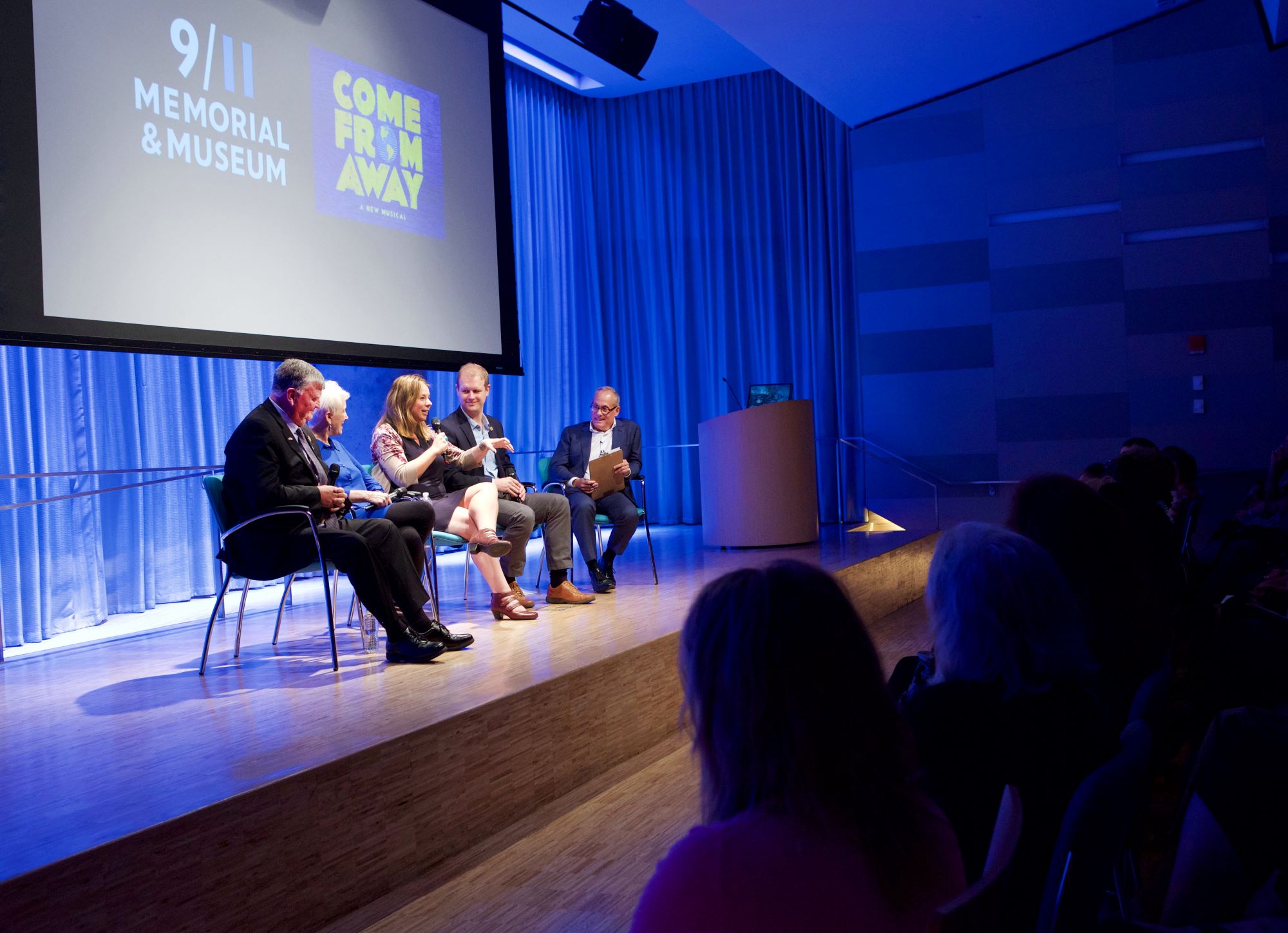 People behind the award-winning new musical “Come From Away” take part in the public program, Come From Away. They include two women and two men. Clifford Chanin, the executive vice president and deputy director for museum programs, sits next to the four attendees. Members of the audience can be seen in the foreground, their profiles bathed in the blue light of the stage. 