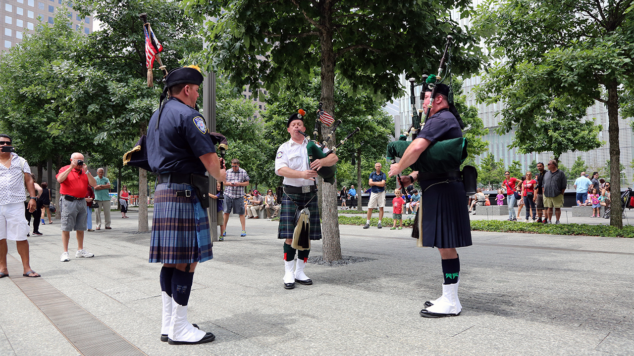 Three men wearing kilts stand in a circle while playing bagpipes on the 9/11 Memorial Plaza in daytime.   Visitors to the Memorial are standing on the periphery, listening to the music.  Two people are taking photos of the bagpipers.