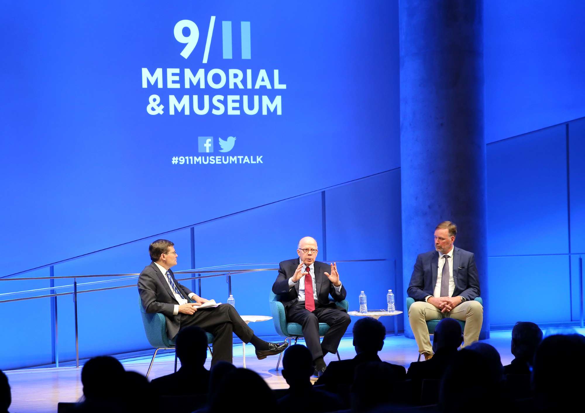 Former Acting CIA Directors John McLaughlin and Michael Morell and former CIA Senior Paramilitary Officer Phil Reilly sit beside each other onstage in this wide angle photo of the Museum Auditorium. McLaughlin is seated between the other two men and gesturing with both hands as he speaks to the audience. Members of the audience are seated in the foreground. Their figures are silhouetted by the bright blue lights behind the stage. The 9/11 Memorial & Museum logo is displayed on a wall above the three partici