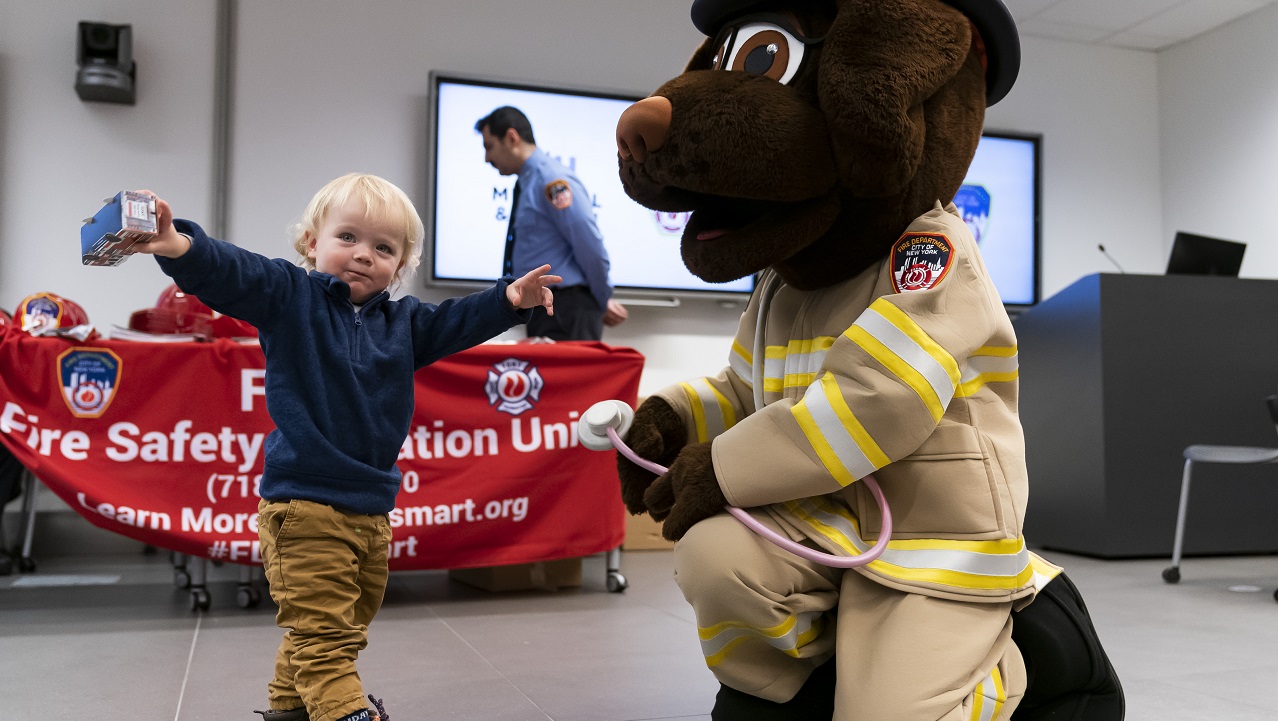A person in a FDNY Fire & Life Safety dog mascot suit greets a young visitor, whose arms are outstretched.
