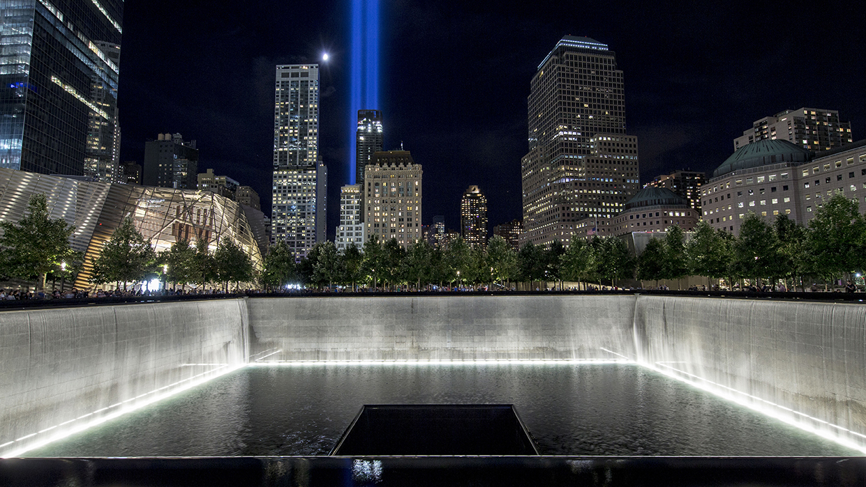 The blue lights from Tribute in Light shine over the illuminated memorial pool at night.