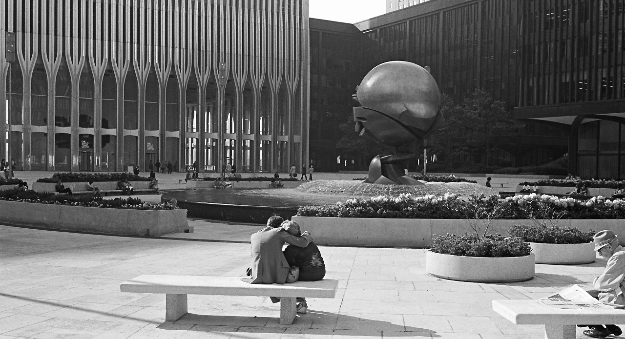 Black and white photo of a man sitting on a white bench with his arm around a woman, facing a spherical metal sculpture at the center. On the left is the ground level view of one of a Twin Tower.