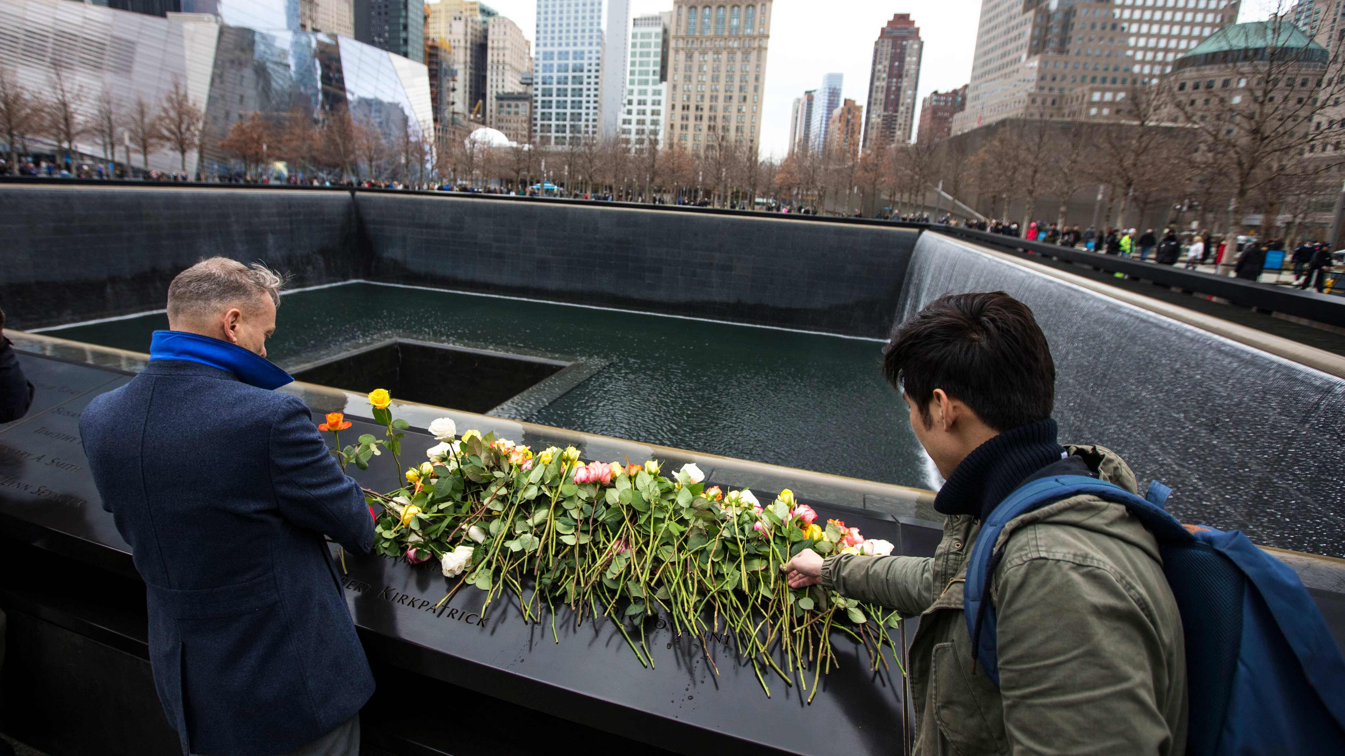 Two people place roses on the dozens of roses already left on the 1993 panel of the 9/11 Memorial.  Behind the panel water flows down the Memorial and into the Memorial Pools.  Lower Manhattan is background beyond the Memorial. 