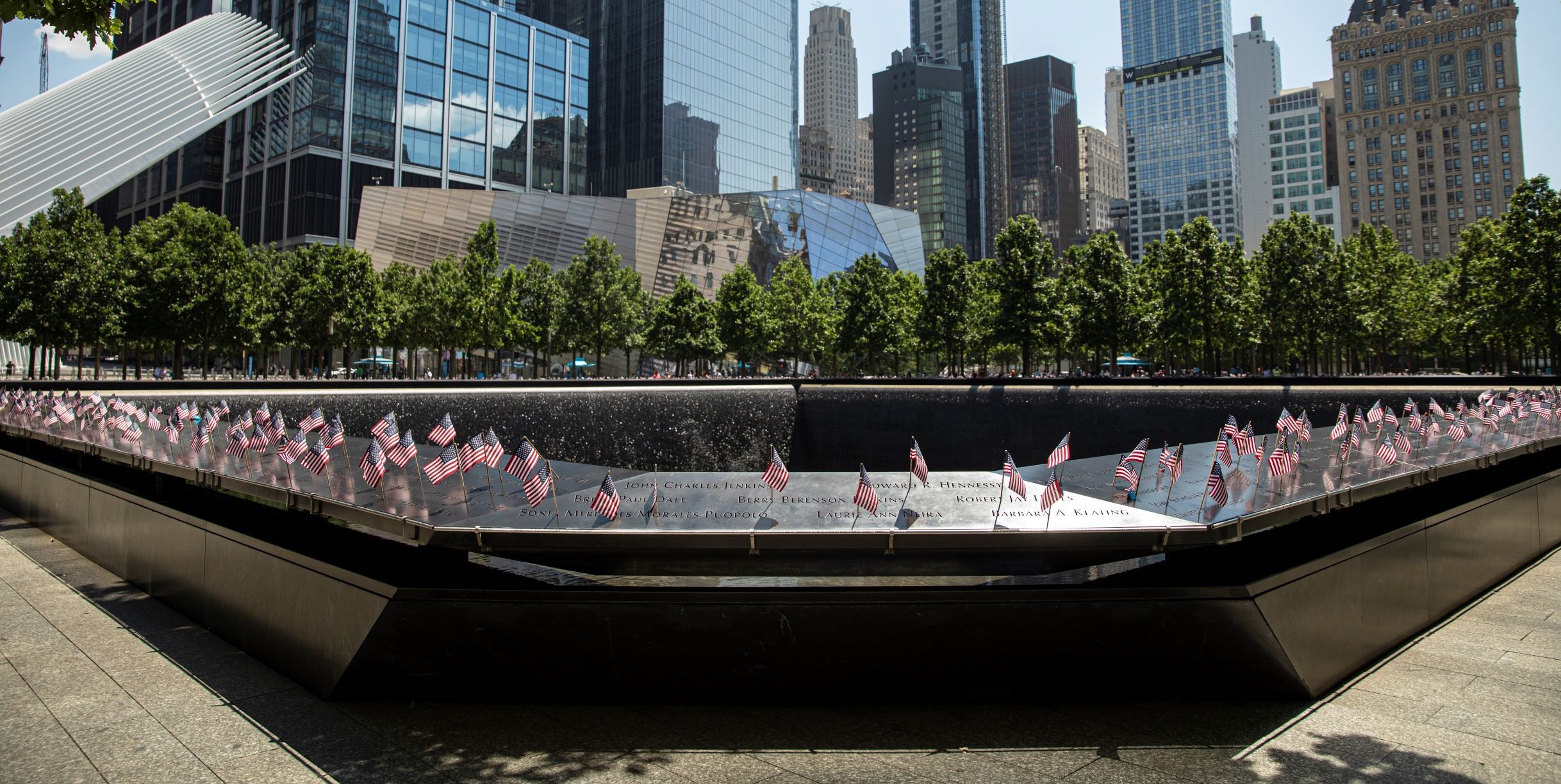 America flags adorn the names on the bronze parapets of the 9/11 Memorial on a bright, sunny day. In the background are full, green trees and the Memorial Museum. 