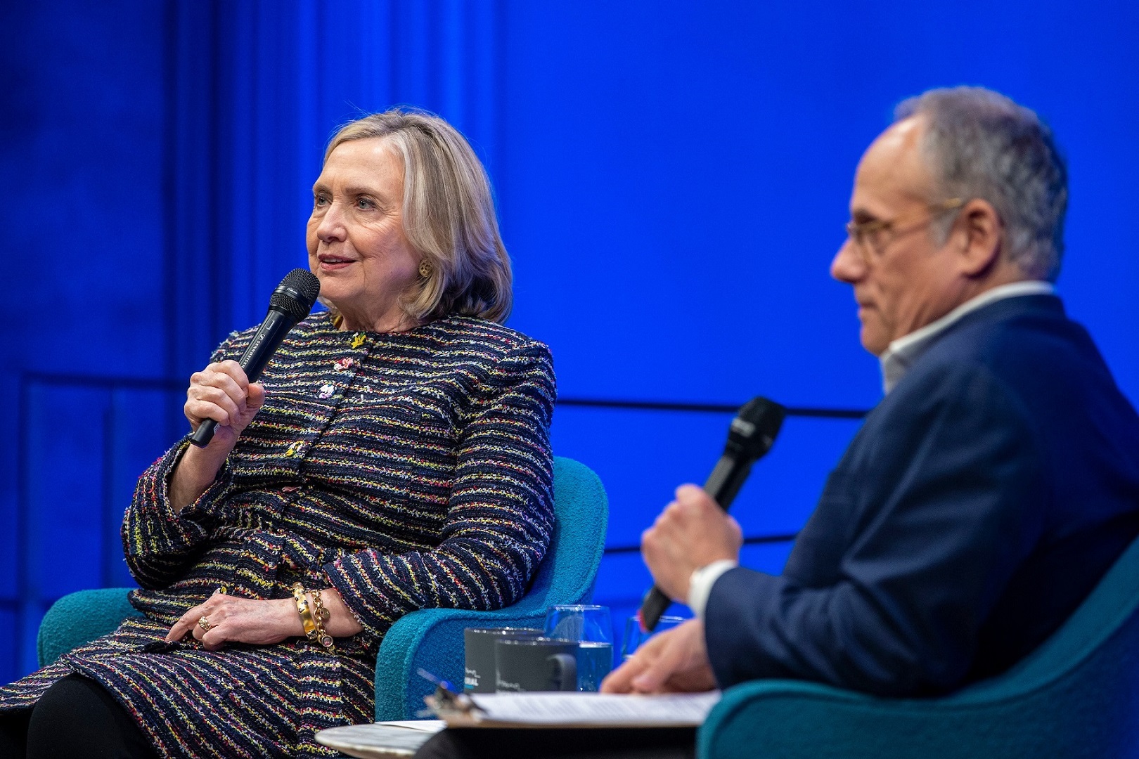 Hillary Rodham Clinton is seated, holding a microphone, to the left of a man in a navy blue suit and glasses, also with a microphone
