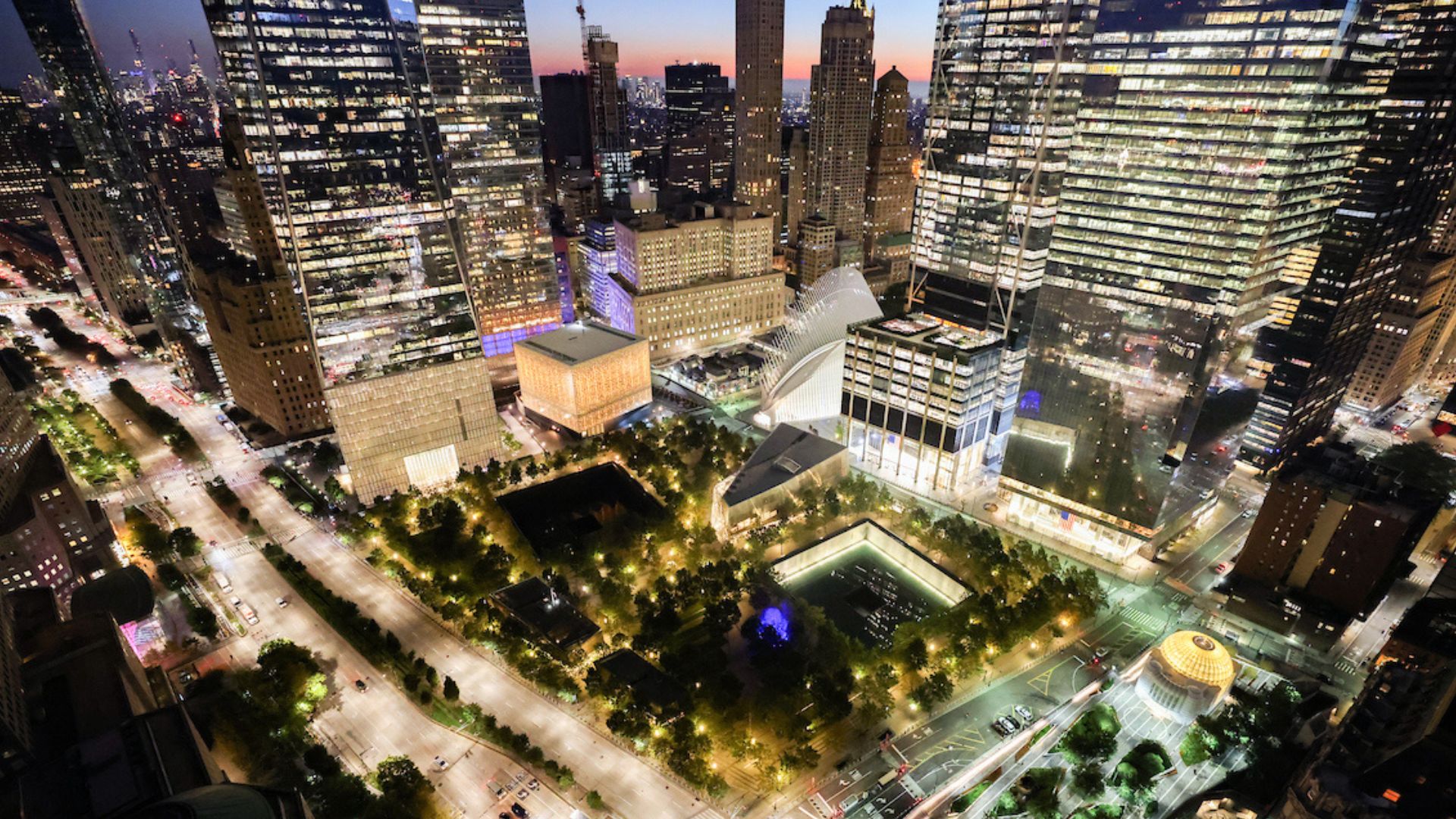 Nighttime aerial view of illuminated Memorial plaza and Museum, framed by (clockwise from left) One World Trade Center, the PAC, the Oculus, the PAC, and St. Nicholas Greek Orthodox Church