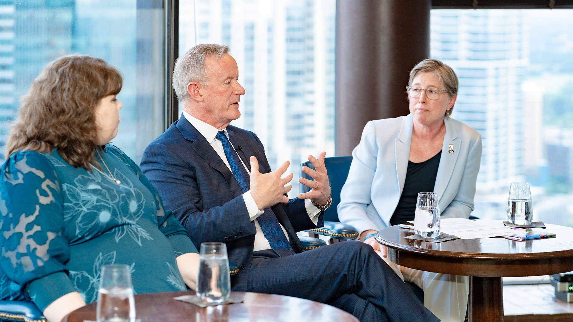 Three speakers, one man and two women, seated in front of a view of the city out of the window behind them