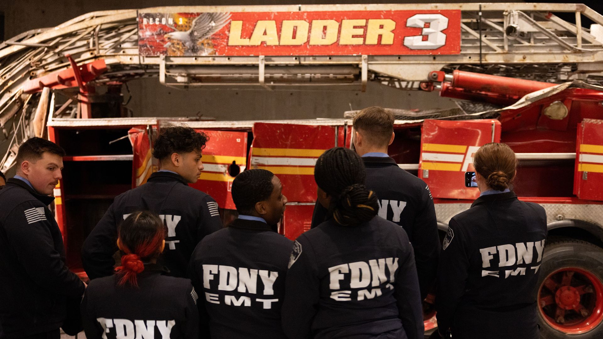 A group of men and woman wearing navy blue jackets that say FDNY EMT on them in white look at the mangled Ladder 3 fire truck