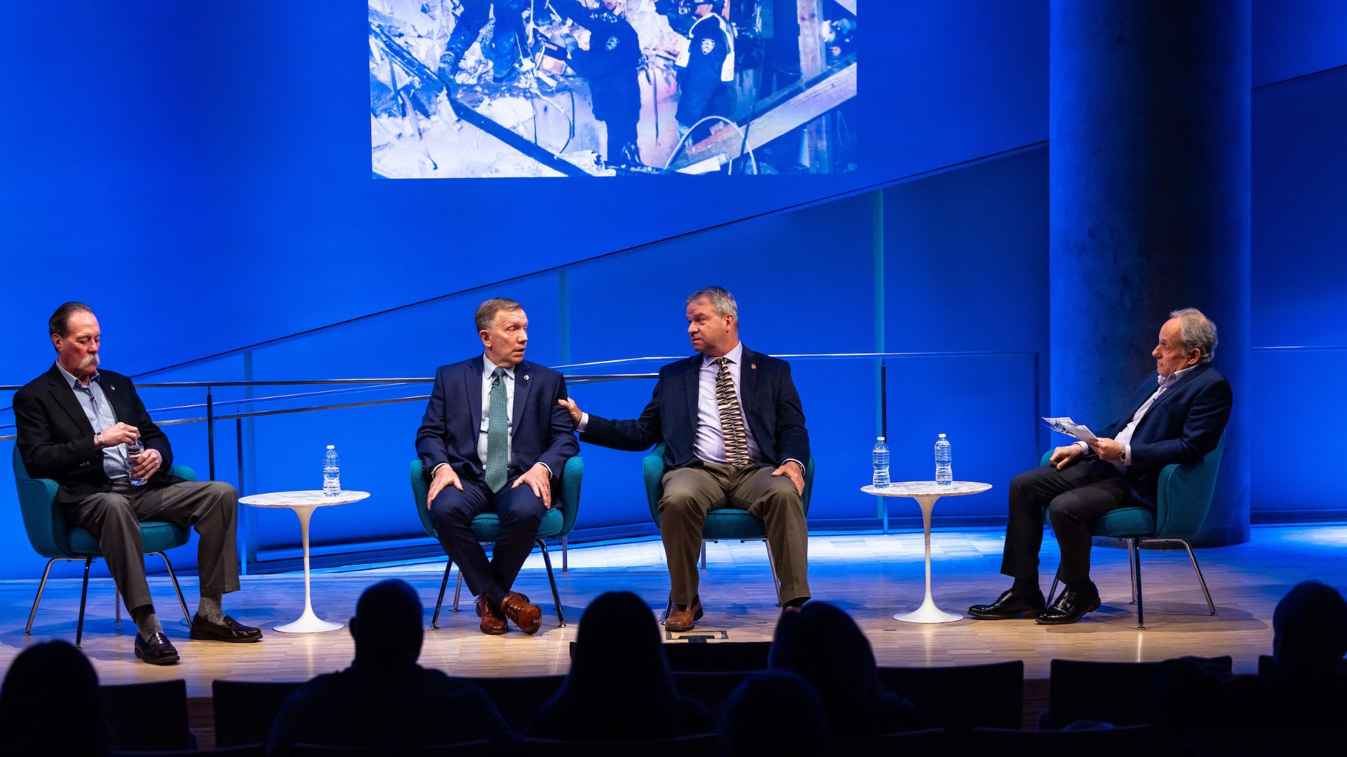 Four panelists sit on a stage with a blue curtain behind it, and a screen showing a snippet of rescue and recovery operations