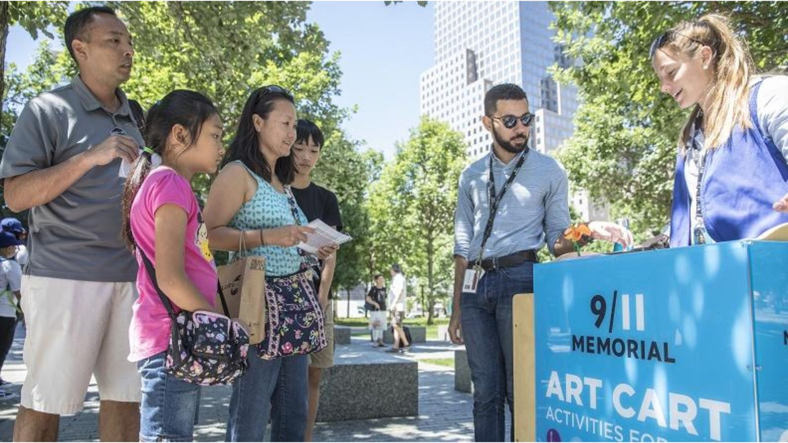On a sunny day outdoors, a group of adults and children watch as a museum staffer stands behind a waist-high cart that says "9/11 Memorial Art Cart Activities."