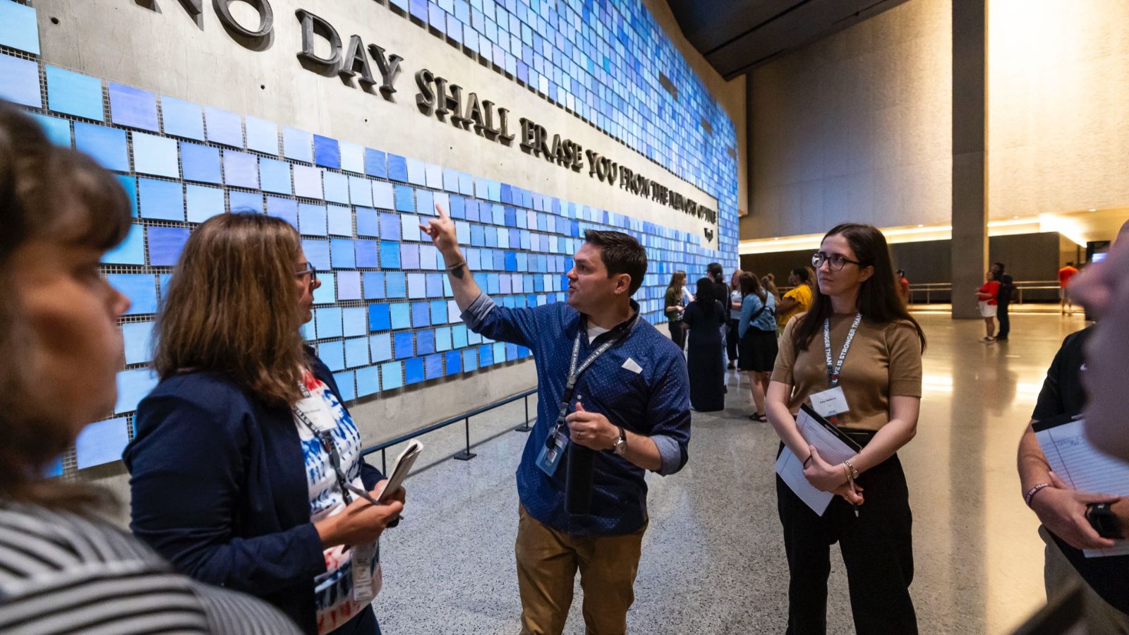 A museum guide points to a wall decorated with blue tiles and the quote "No day shall erase you from the memory of time" as a small group of adults looks on.