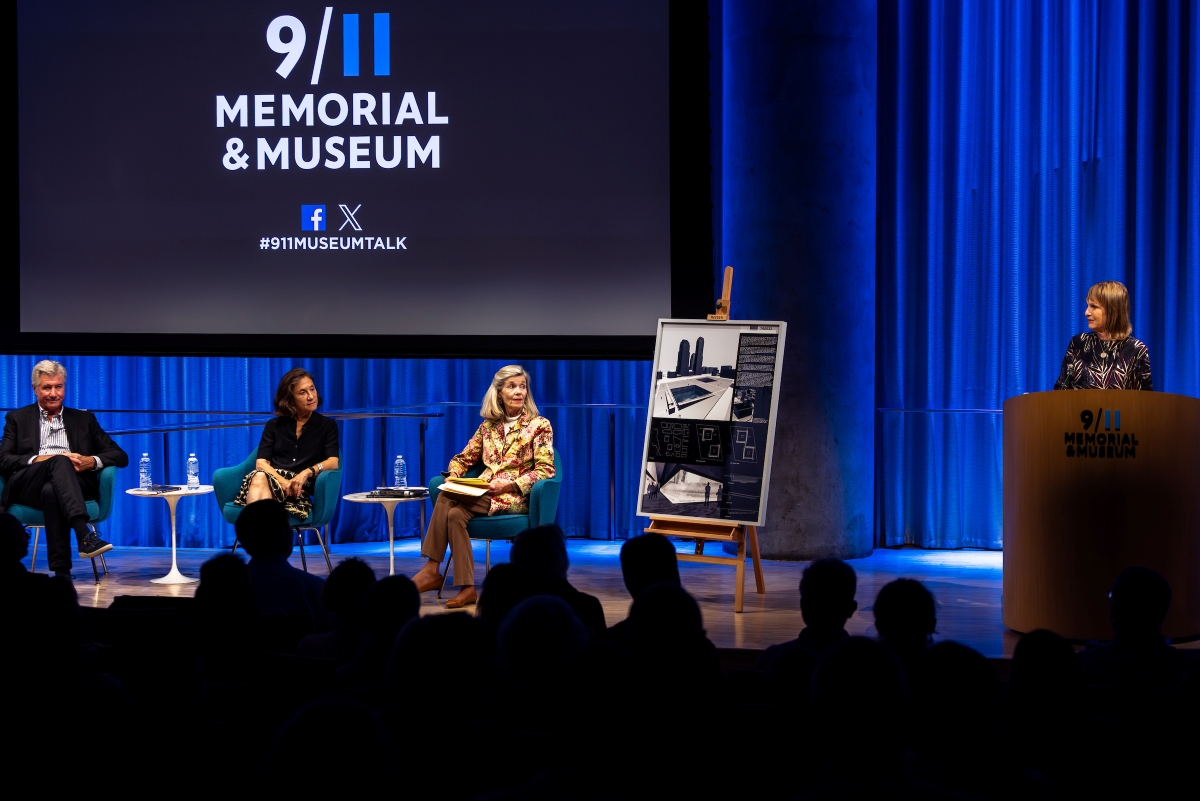 Speakers sit on stage as they are introduced by a woman at the podium.