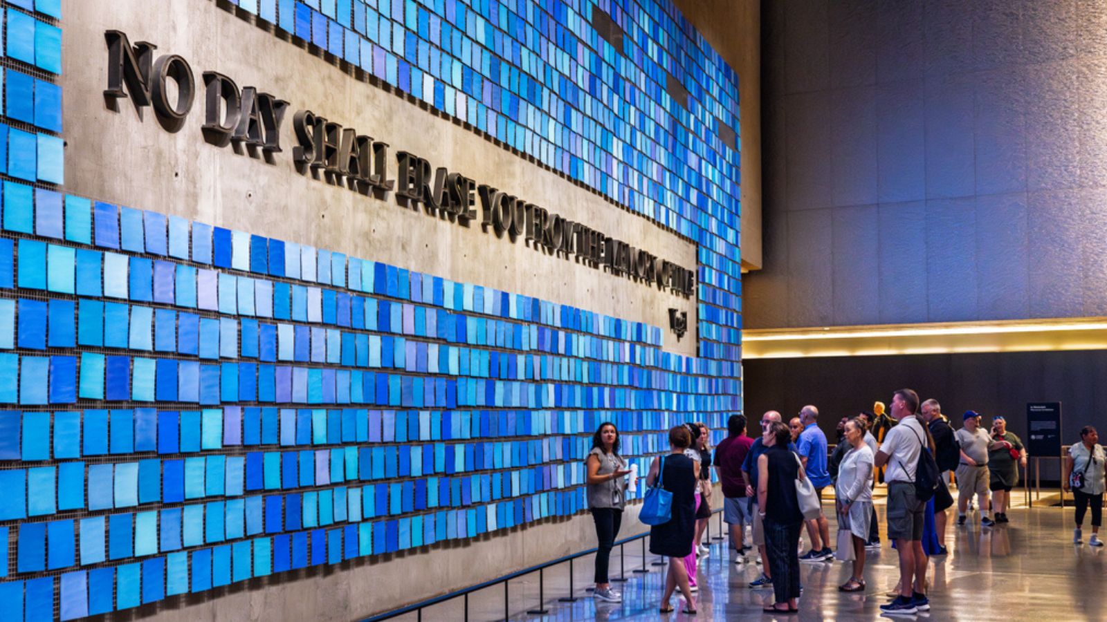 A group of visitors looks at a wall of tiles in different shades of blue, with the quote No Day Shall Erase You from the Memory of Time visible