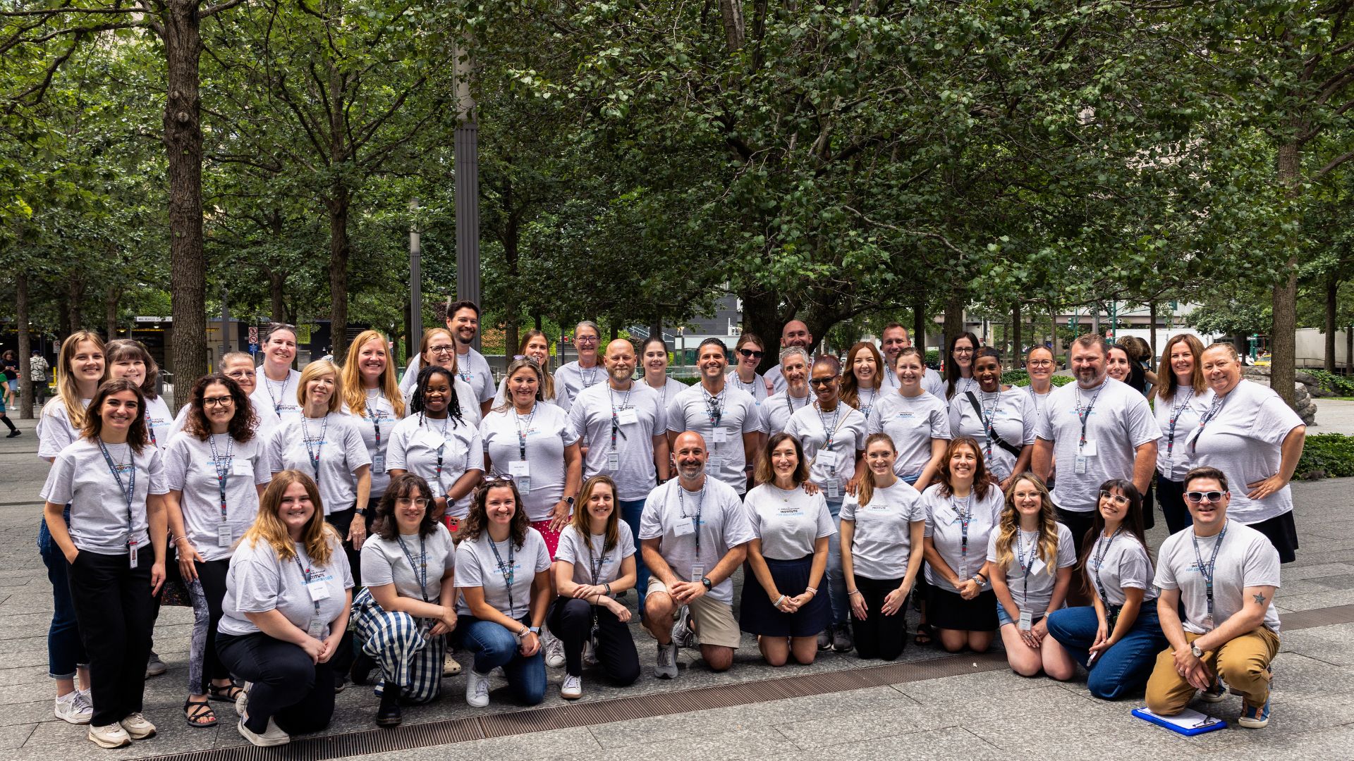 A group of adults in gray tee shirts pose, smiling, in three rows on the Memorial