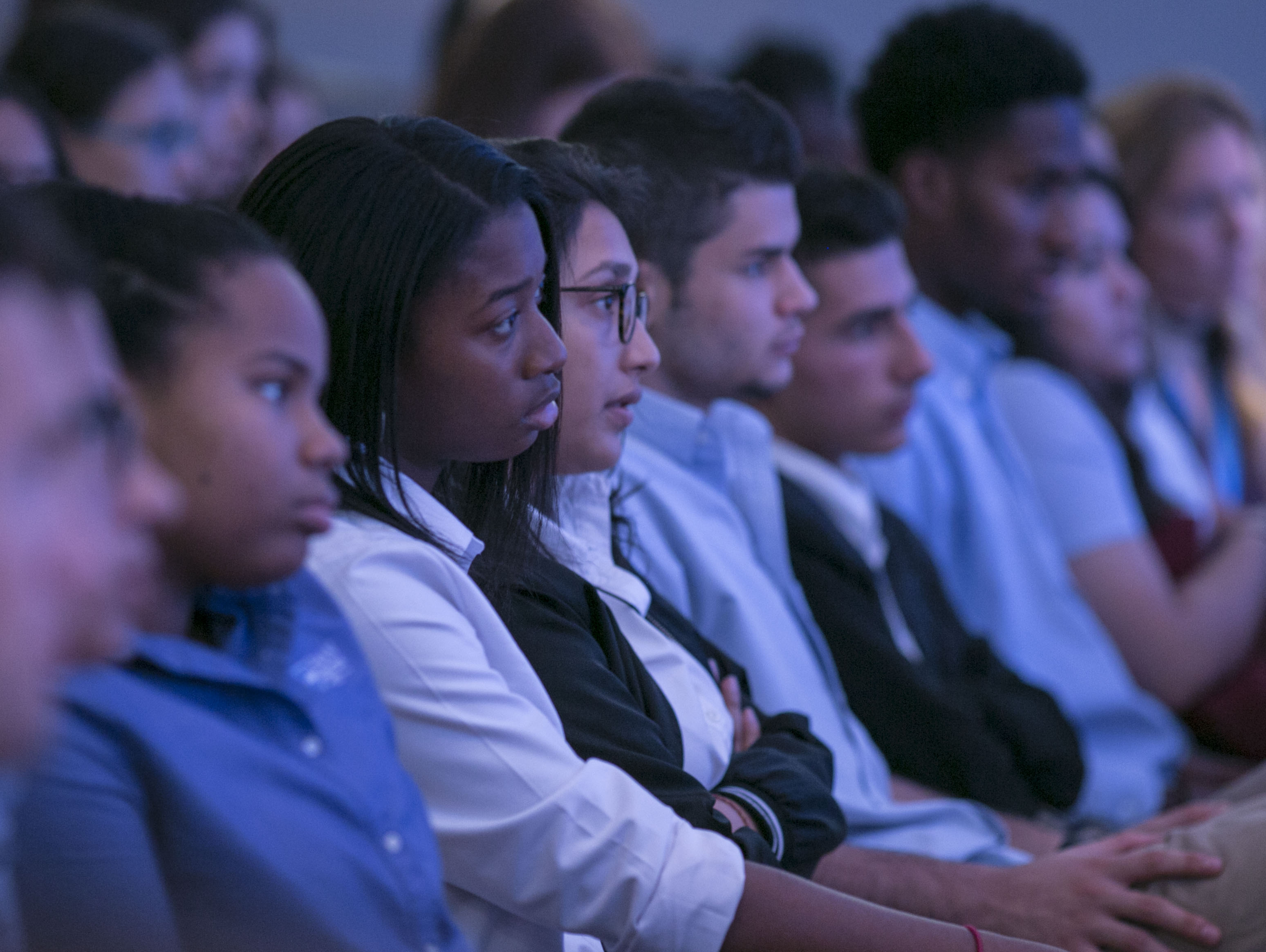 Upper elementary aged students sit in a row listening to a speaker