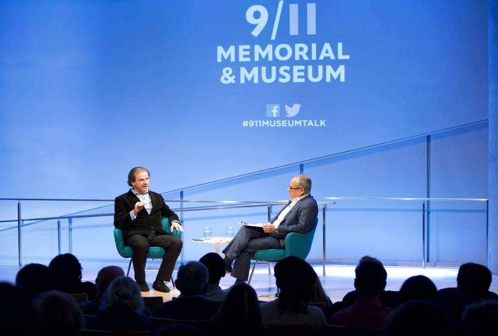 New York Times best-selling author and CNN National Security Analyst Peter Bergen sits onstage with Clifford Chanin, the executive vice president and deputy director for museum programs, during a public program at the Museum Auditorium. Members of the audience are silhouetted in the foreground. The 9/11 Memorial & Museum logo is projected on a wall above them.