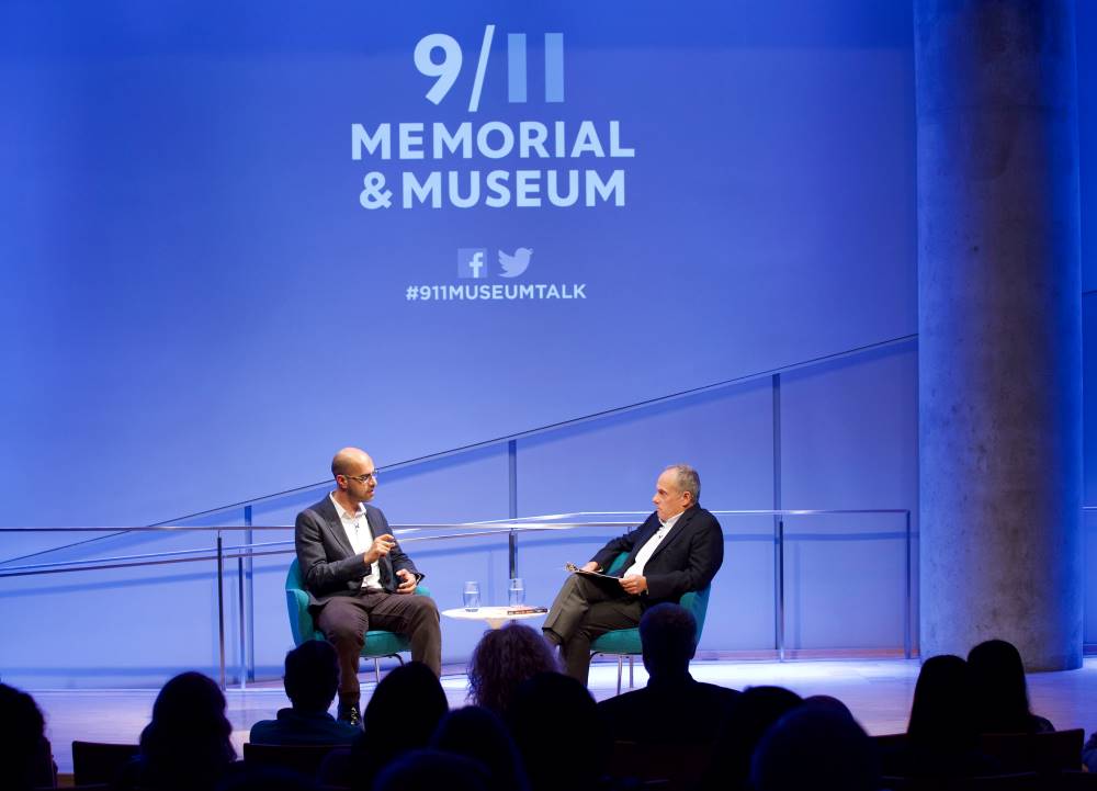 Author and news commentator Haroon Moghul speaks onstage as he takes part in the public program, How to Be a Muslim, in this photo from the audience. Clifford Chanin, the executive vice president and deputy director for museum programs, looks on as he sits next to Moghul. Audience members in the foreground are silhouetted by the lights onstage.