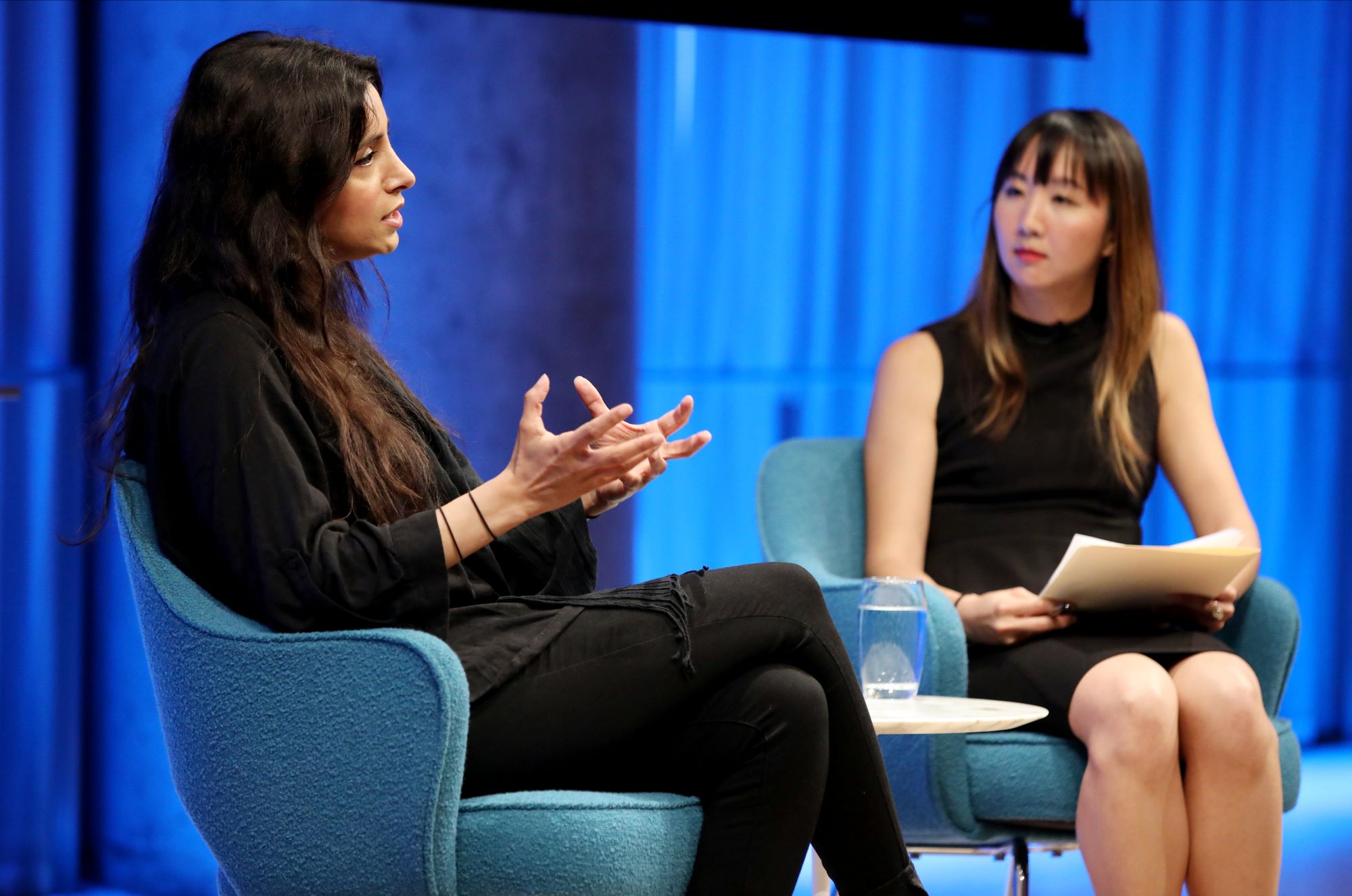 Emmy Award–winning documentarian Deeyah Khan gestures with both of her hands as she speaks while looking towards the audience, which is out of view, in the Museum Auditorium. The woman hosting the event sits to the left of Khan. She listens and looks at Khan.