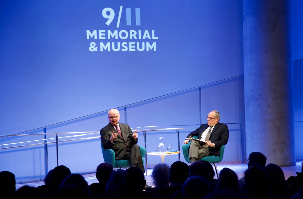 Former CIA director and retired U.S. Air Force Gen. Michael Hayden speaks onstage as part of the public program, General Michael Hayden on the War on Terror. Clifford Chanin, the executive vice president and deputy director for museum programs, watches from the stage as he holds a clipboard. The silhouettes of audience members are in the foreground. The logo of the 9/11 Memorial & Museum is projected above Hayden and Chanin.