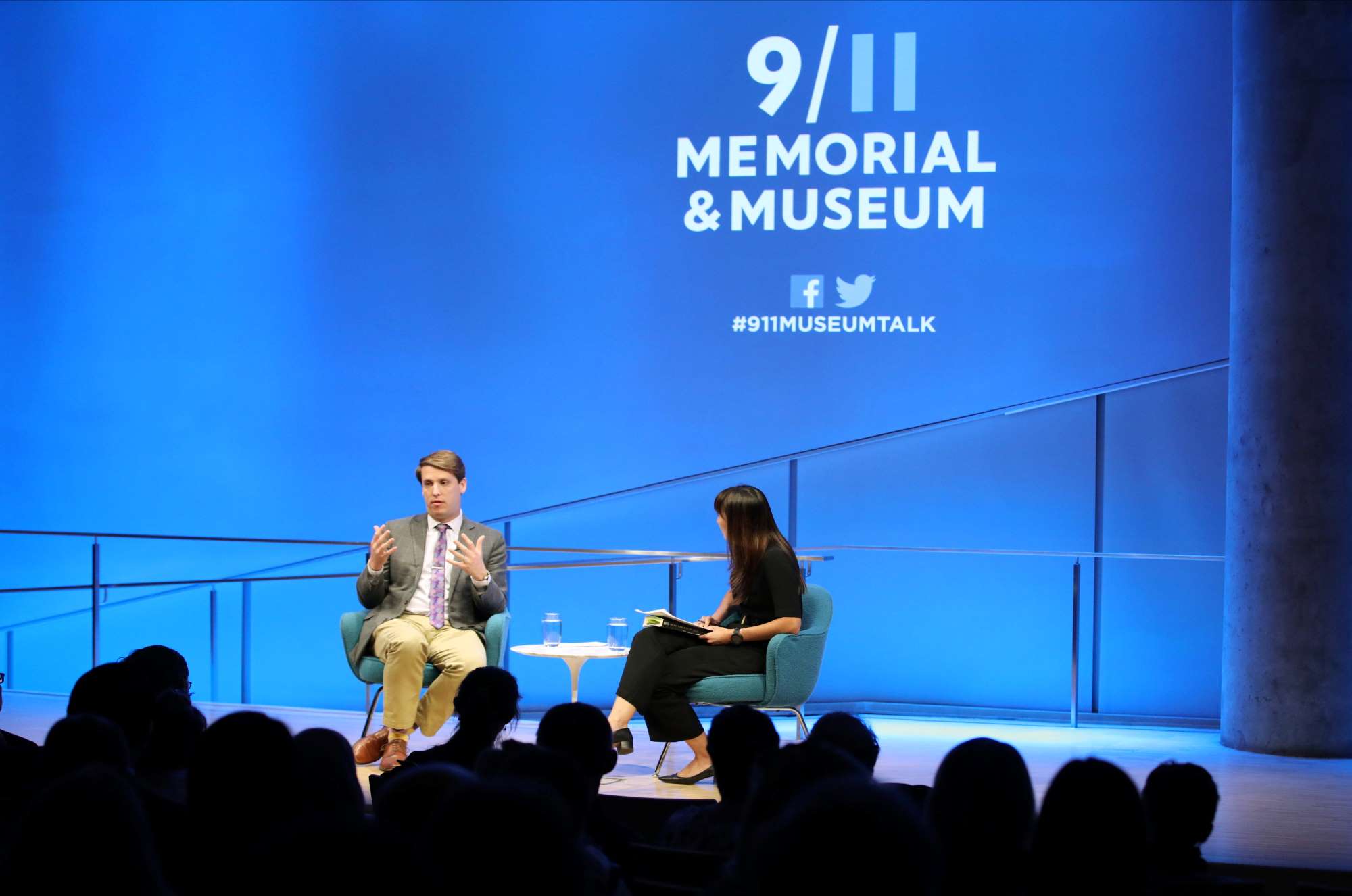 This wide-angle photo of the Museum Auditorium shows journalist and historian Garrett Graff seated onstage with the woman who is hosting the public program “Only Plane in the Sky. Graff is gesturing with both hands as he looks towards the audience, whose heads are silhouetted in the foreground. A wall behind Graff is lit blue and features the logo of the 9/11 Memorial & Museum.