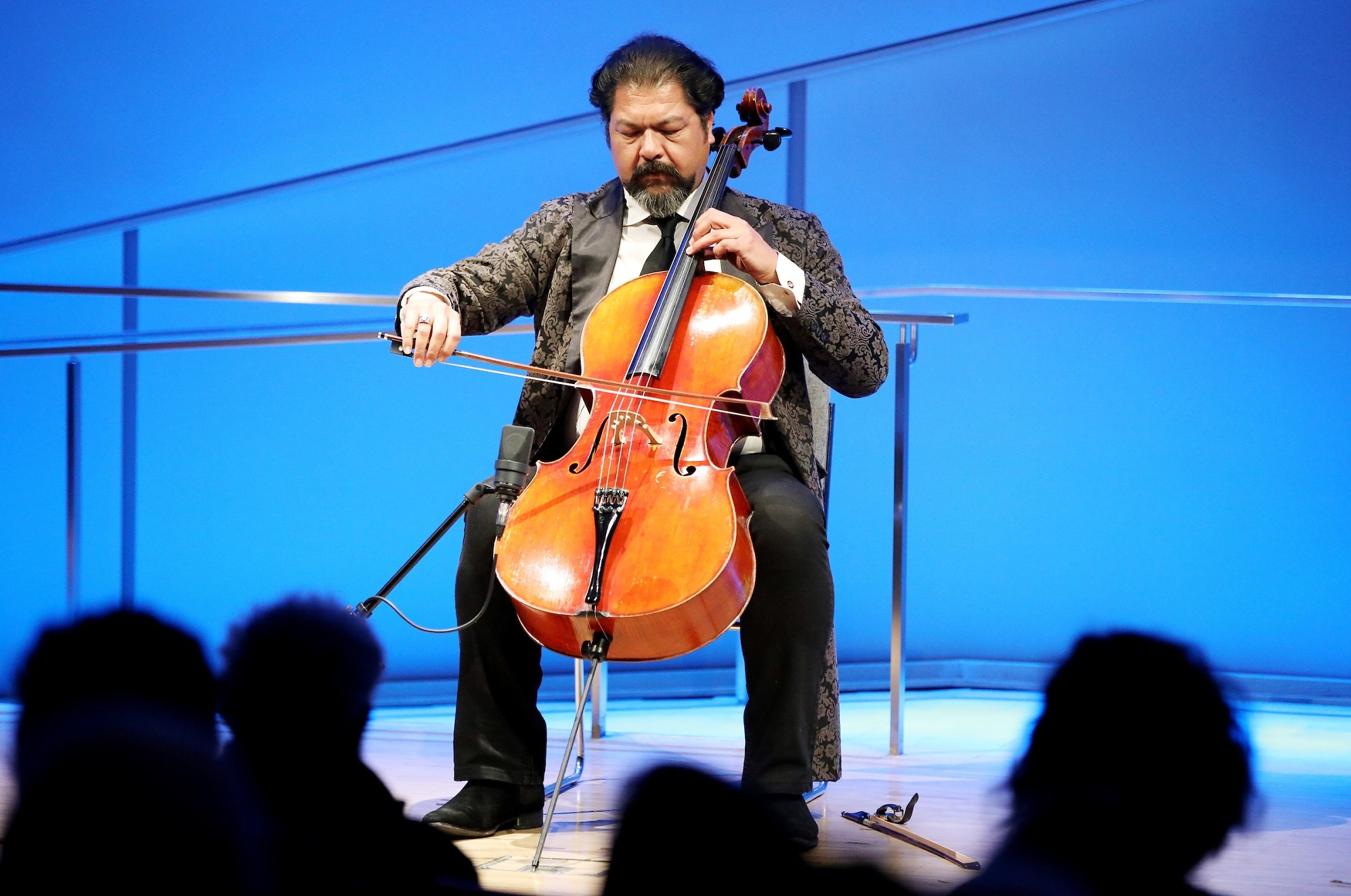 Cellist Karim Wasfi performs while seated onstage at the Museum auditorium. His eyes are closed as if he’s concentrating. His right hand grasps the bow as his left hand moves along the strings. His bright orange-brown cello contrasts with the blue lights of the stage. The silhouettes of several audience members are visible in the foreground.