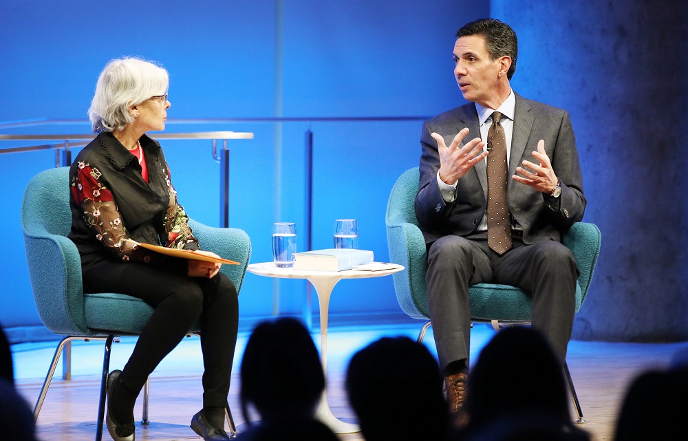 In this photo taken from behind the audience, Boston Globe reporter Mitchell Zuckoff speaks onstage while gesturing with both of his hands. He is speaking to a woman who is hosting the public program at the Museum Auditorium. She is seated next to him and holding a manila envelope. The silhouetted heads of several audience members are visible in the foreground.