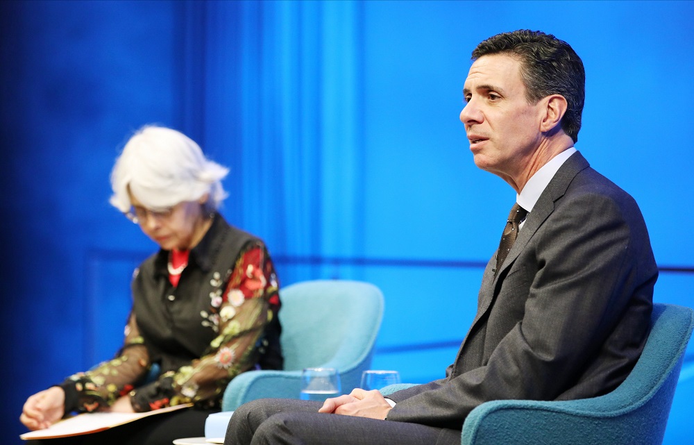 Boston Globe reporter Mitchell Zuckoff looks towards the audience with his hands in his lap while speaking onstage at the Museum Auditorium. A woman who is hosting the event looks down at a piece of paper as she sits to his right.