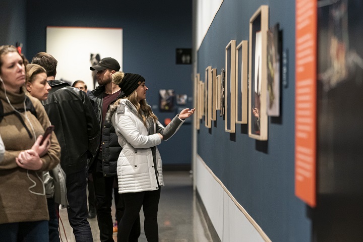 Museumgoers in winter coats stand in front of a blue wall, looking at framed photos in an exhibition. Some visitors are wearing audio guide headsets.