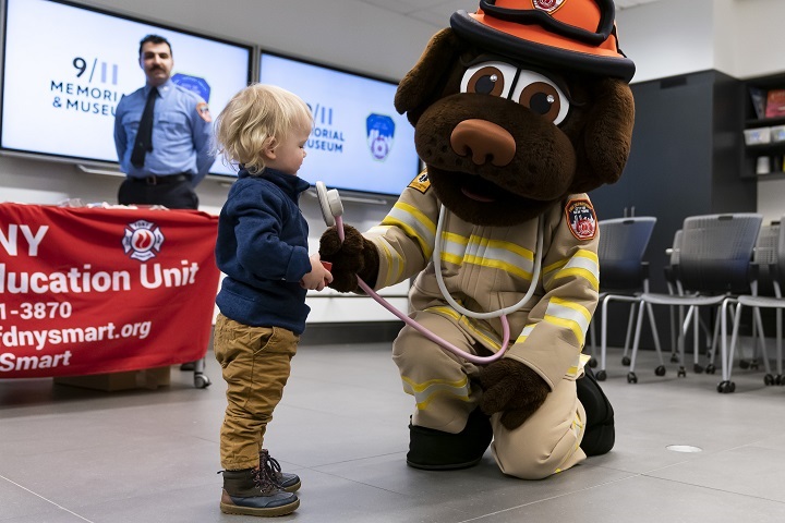 A man in a dog suit kneels and presses a plush stethoscope to a blond toddler's chest.