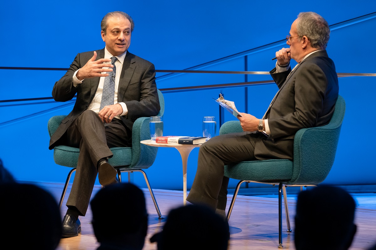 In this view from the audience, Preet Bharara, the former U.S. attorney for the Southern District of New York, speaks onstage as he gestures with his right hand. Moderator Clifford Chanin sits to Bharara’s left holding a clipboard and putting a pen to his chin. A small white table with two glasses of water on it is between them. The wall behind them is lit blue. The silhouettes of audience members are in the foreground.