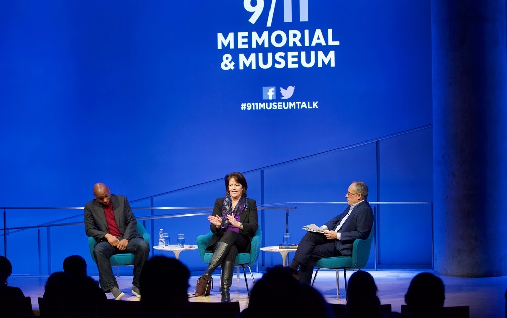 Three public program participants sit on a blue-lit auditorium stage. “9/11 Memorial & Museum #museumtalk” is projected onto the wall behind them. To the far left, a man in a gray blazer, red sweater, and jeans looks down at his feet, while a woman in the center sits with crossed legs, gestures with her hands, and addresses the audience. A man with a clipboard in his lap watches her and listens. The heads of the audience members appear in the foreground in silhouette.