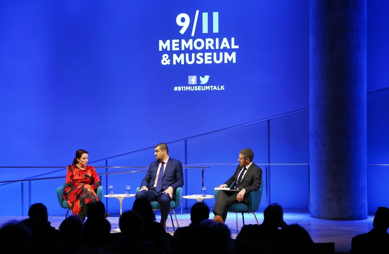 American Enterprise Institute scholar Karen E. Young, journalist Hassan Hassan, and the 9/11 Memorial & Museum’s Noah Rauch sit onstage during a public program at the Museum’s Auditorium. In this view from the audience, Young is speaking as Hassan and Rauch look towards her. About a dozen silhouetted audience members are in the foreground. Blue lights create a blue aura behind the participants onstage.