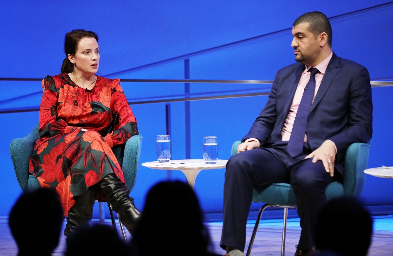 American Enterprise Institute scholar Karen E. Young sits with her legs crossed as she speaks onstage at the Museum Auditorium. A dress she is wearing with bright red flowers contrasts with the blue lights of the stage. To her left is journalist Hassan Hassan, who is listening with his hands on his legs. The heads of several audience members are silhouetted and out of focus in the foreground.