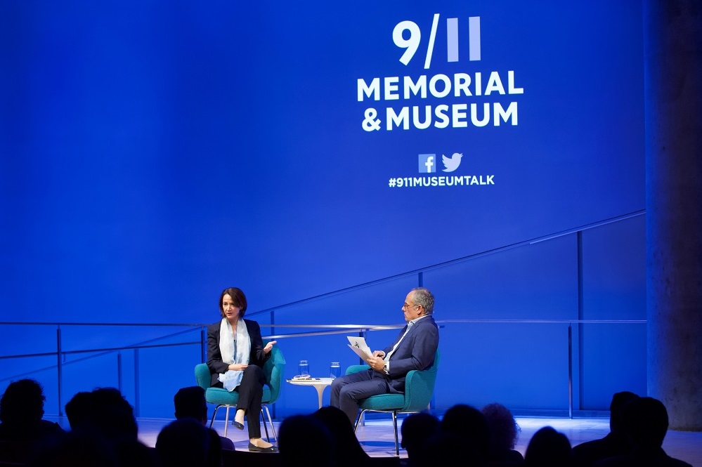 In this wide-angle photograph, a woman in a gauzy scarf and a navy-blue suit sits on a blue-lit auditorium stage and gestures with her hands while she looks out into the audience. A man sits beside her on stage and holds a clipboard in one hand while listening intently. “9/11 Memorial & Museum #museumtalk” is projected onto the stage behind them. The heads of the audience appear in the foreground in silhouette.