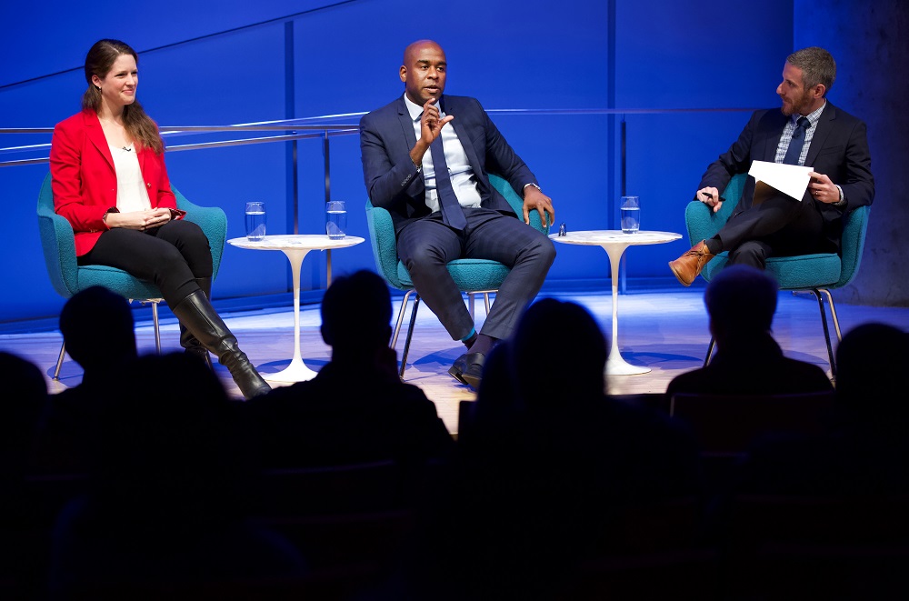 Three public program participants sit on a blue-lit auditorium stage. On the far left, a woman in a red blazer smiles looks off into the audience. In the center, a man gestures with his hands and out into the audience. The moderator sits crossed legged with a clipboard in his lap and looks at the man speaking. The heads of the audience members appear in the foreground in silhouette.