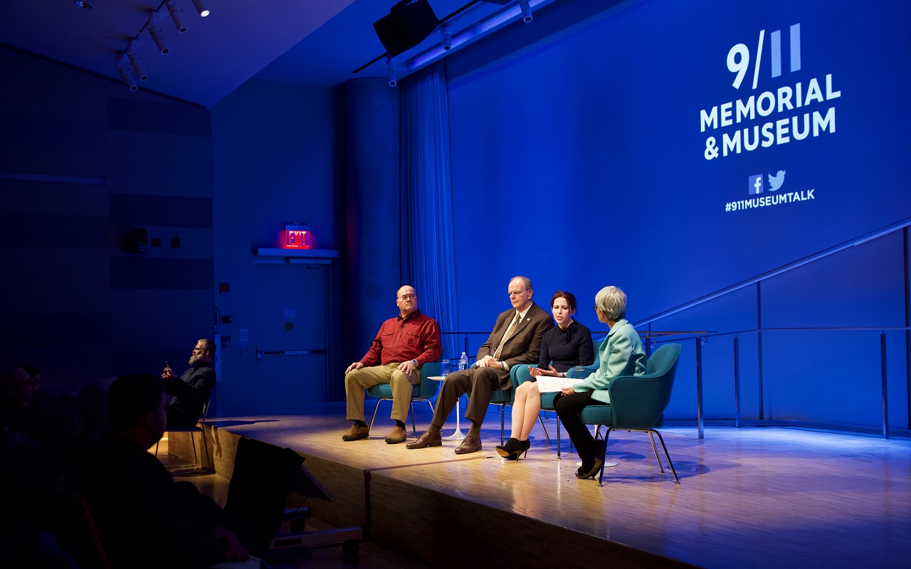 A public program moderator and three guests take part in a public program onstage at the Museum’s Auditorium in this view from the audience. The stage lights are shining on the four of them and the rest of the auditorium, including the audience, is in the dark. The guests include, from left to right, Kahnawake Council Chief Lindsay LeBorgne, Local 40 Business Manager Robert Walsh, and artist Melissa Cacciola, who is speaking beside the moderator. A sign language interpreter is seated in the distance.