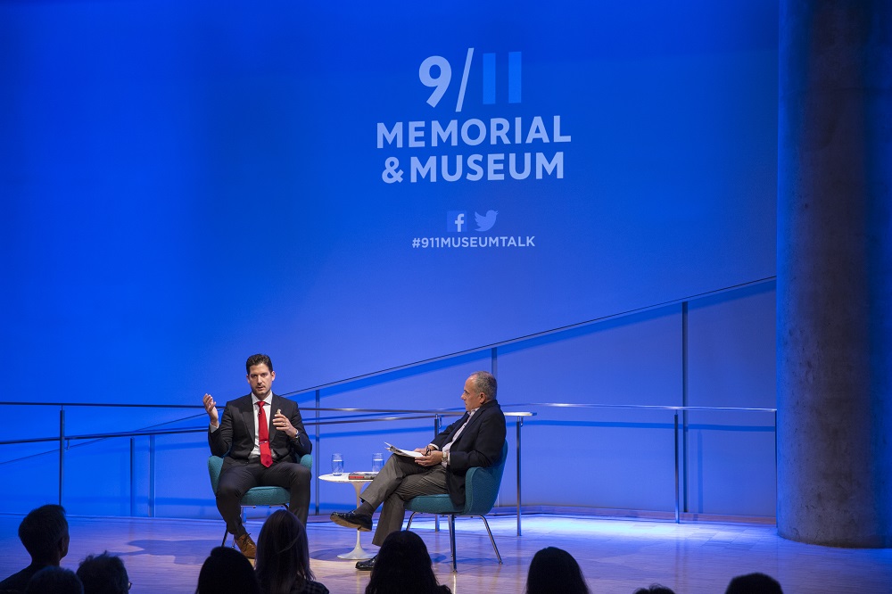 In this photograph, two men sit on a blue-lit auditorium stage, with “9/11 Memorial & Museum #911museumtalk” projected onto the wall behind them. One man in a suit and red tie gestures with his hands toward the audience, while the other man sits with a clipboard in his lap. The audience appears in silhouette in the foreground of the photograph.