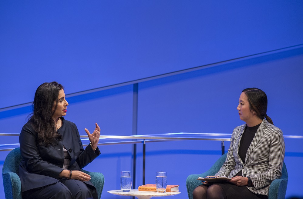 In this photograph, two women sit on a blue-lit auditorium stage. One woman in a dark gray suit gestures with her hands while another woman in a light gray blazer listens.