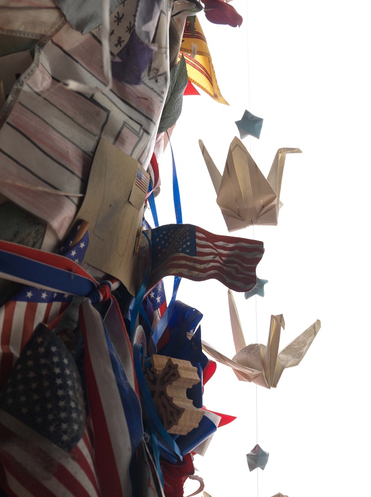 Detailed shot of Lady Liberty sculpture shows a juxtaposition of mini American flags and a small chain of two white papercranes hanging off of the statue's side