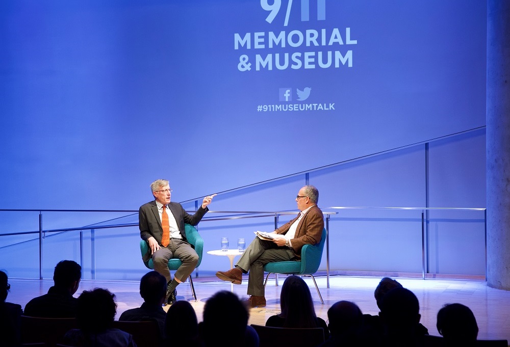 Two men sit on a blue auditorium stage with “9/11 Memorial & Museum #museumtalk” projected onto the wall behind them. One man in a suit and orange tie points with his left hand as he speaks. The other man sits with a clipboard in his lap and listens. The heads of the audience members appear in silhouette in the foreground of the photograph.