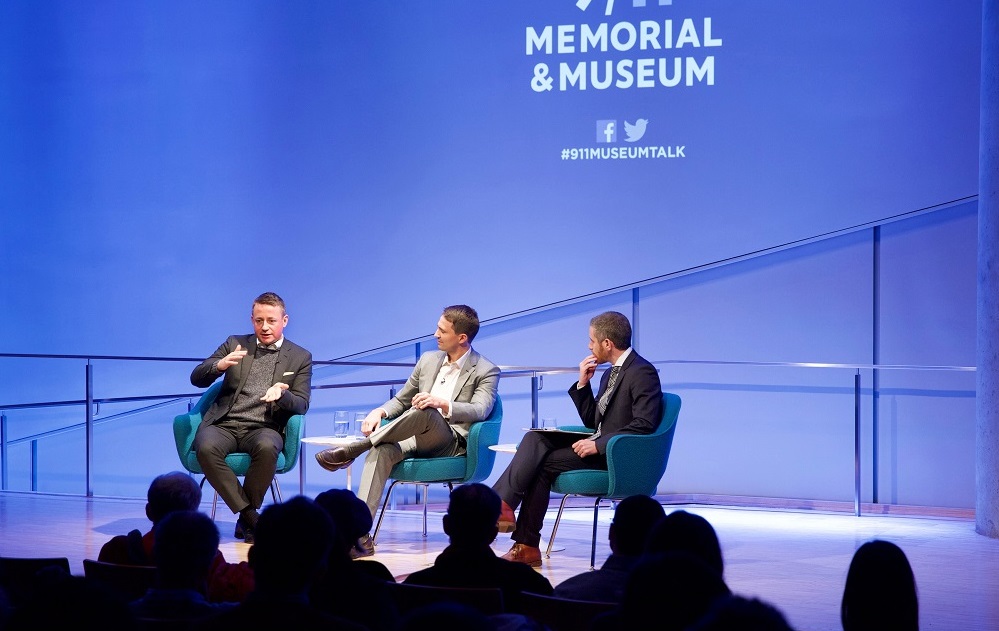 In this wide-angle photograph, three men in suits sit on a blue-lit auditorium stage with “9/11 Memorial & Museum #911museumtalk” projected onto the wall behind them. A man in a charcoal gray suit speaks and gestures with his mans while the other two men look on. The heads of the audience members appear in the foreground in silhouette.